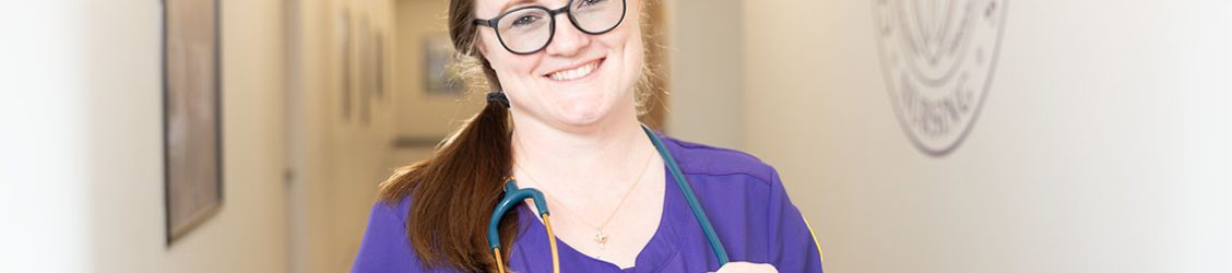 Smiling CTX nursing student in purple scrubs stands in a hallway